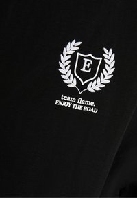 Musta puuvillapaita, jossa on valkoinen brodeerattu emblem, jossa on kilpi ja laakerinlehdet. Tekstissä lukee "team flame. NAUTI TIESTÄ."