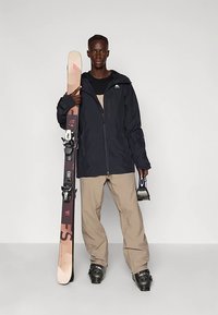 Črna vodoodporna smučarska jakna, bež hlače za sneg, v roki drzijo rjavo desko za deskanje na snegu z vezmi. Gladke teksture, praktičen kroj za zimske športe.