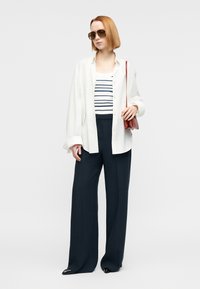 Λευκό πουκάμισο με κουμπιά πάνω από ριγέ αμάνικη μπλούζα, συνδυασμένο με ναυτικό μπλε παντελόνι με φαρδιά πόδια. Αξεσουάρ περιλαμβάνουν καφέ γυαλιά ηλίου και κόκκινη τσάντα.