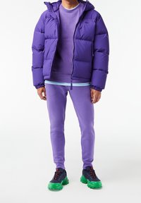 Chaqueta de plumas morada con capucha, combinada con un sudadera morada más clara, joggers a juego y zapatillas multicolor con suelas verdes.