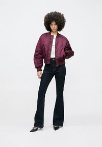 Chaqueta bomber color burdeos con puños acanalados, combinada con vaqueros acampanados oscuros de talle alto y tacones negros puntiagudos. Top blanco visible debajo.