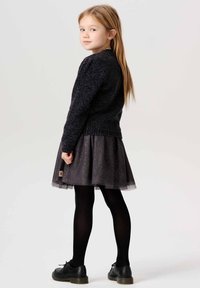 Zwarte gebreide trui gecombineerd met een donkergrijze tule rok met sprankels. Meisje draagt zwarte panty's en schoenen, wat een casual outfit laat zien.