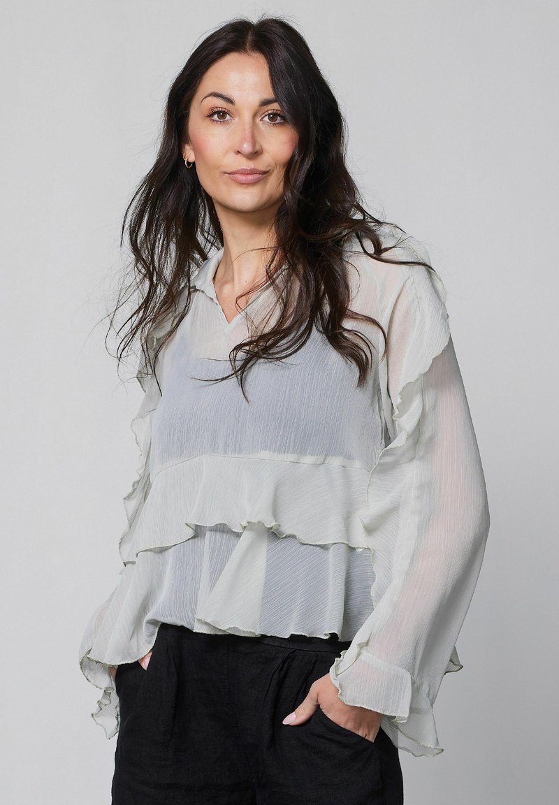 Femme aux longs cheveux foncés portant un chemisier blanc transparent à volants et un pantalon noir, debout les mains dans les poches devant un fond uni.