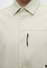 Chemise beige clair à boutons avec col et détail boucle en corde noire sur la poche poitrine supérieure.