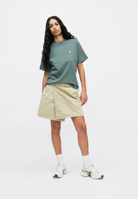 Πράσινο βαμβακερό T-shirt με μικρό κίτρινο λογότυπο, σε συνδυασμό με μπεζ φούστα τύπου περιτυλίγματος. Το μοντέλο φοράει λευκά και πράσινα αθλητικά παπούτσια και λευκές κάλτσες.