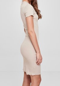 Femme portant une robe beige ajustée à manches courtes avec des bords volants, se tenant en profil latéral contre un fond clair et uni.