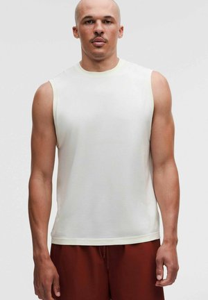 Uomo con testa rasata che indossa una maglia bianca senza maniche e pantaloni rosso scuro, in piedi davanti a uno sfondo chiaro e uniforme.
