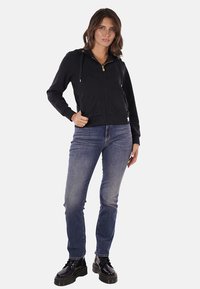 Felpa nera con zip e cappuccio, dettagli dorati e cordini. Indossata sopra jeans blu e stivali neri stringati. Tessuto lisci.