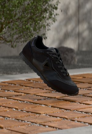 Czarny sneaker z logo po boku wszytym nad brukiem z cegieł i kamieni, na rozmytym tle z drzewem i ścianą.