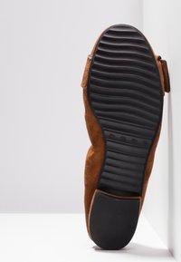 Chaussure en daim marron avec semelle en caoutchouc noire présentant un motif ondulé et un talon solide, placée en position verticale contre une surface et un mur blancs.