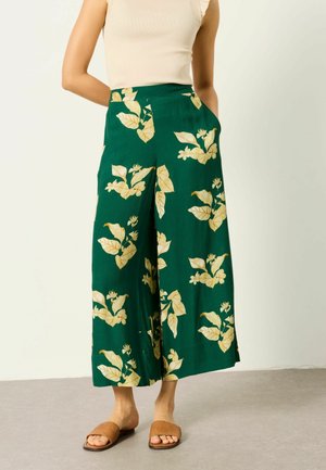 Woman wearing green wide-leg pants with yellow floral print, beige sleeveless top, and brown woven slide sandals, standing with hands in pockets.