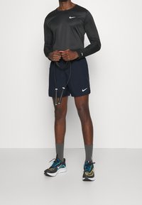Camisa negra de manga larga y pantalones cortos azul marino con los logos del swoosh de Nike, calcetines grises y zapatillas deportivas negras con acentos azules y amarillos; sosteniendo una cuerda para saltar.