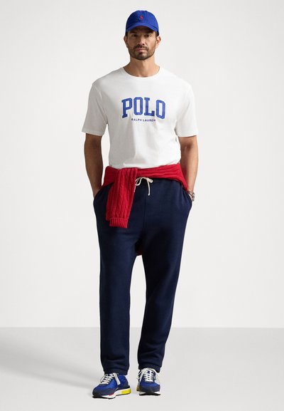 T-shirt blanc avec logo "POLO" bleu, pull en maille rouge noué autour de la taille, pantalon de jogging navy et baskets bleues avec accents multicolores.
