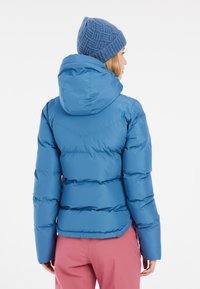 Blaue Pufferjacke mit Kapuze, ausgestattet mit einem gesteppten Design und glatter Textur. Über rosa Hosen getragen, die elastische Bündchen und Saum zeigen.