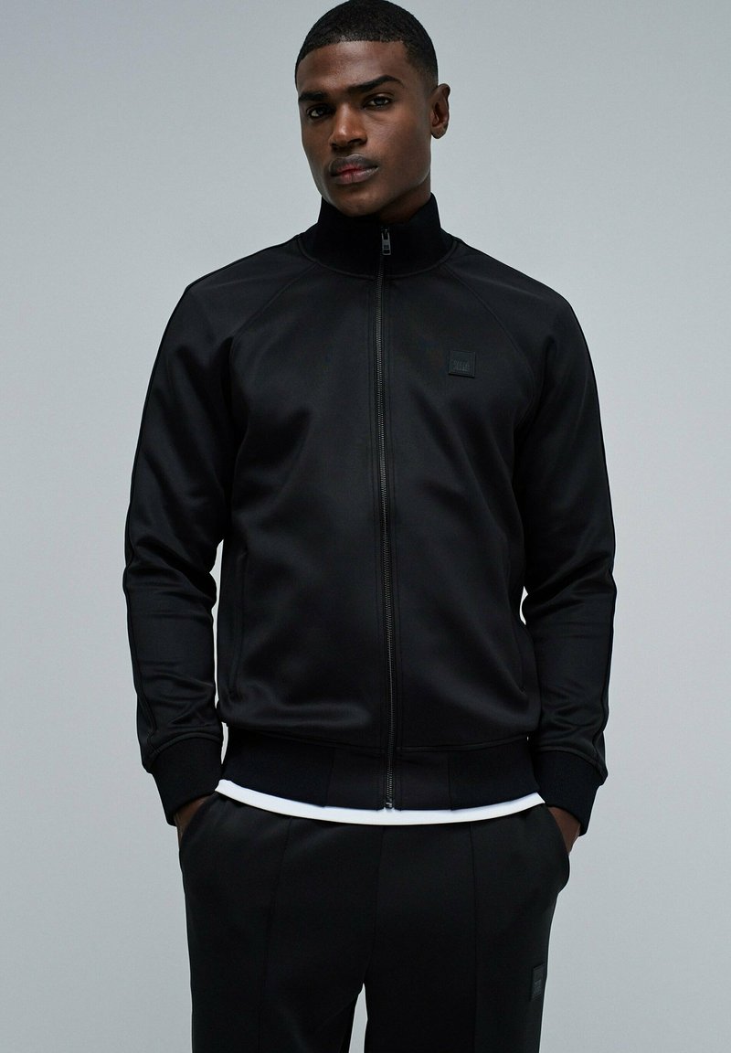 Chaqueta negra con cremallera y alto cuello, fabricada con tejido suave. Presenta puños y dobladillo acanalados, con un pequeño logo en el pecho.