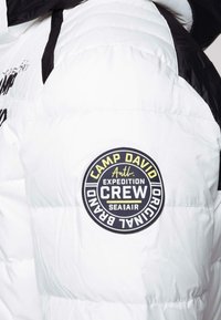 Giacca bianca imbottita con accenti neri; presenta una toppa rotonda sulla spalla con la scritta "Camp David Expedition Crew" in testo giallo.