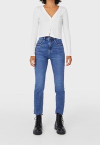 Femme portant un jean taille haute bleu, un cardigan court à manches longues blanc et des bottes noires à lacets, se tenant contre un fond clair et uni.