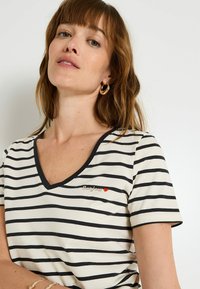 Camiseta de manga corta a rayas en crema y negro. Presenta un escote en forma de V, texto bordado que dice "Bonjour" y un pequeño corazón rojo como acento.