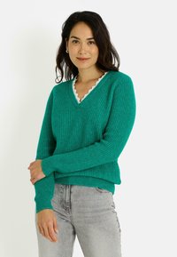 Suéter verde de punto con cuello en V y escote blanco con borde festoneado. Mangas largas y puños acanalados, combinado con vaqueros gris claro.