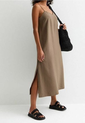 Vrouw met een mouwloze bruine midikleding met een zijsplit, zwarte sandalen en een zwarte gebreide schoudertas in haar hand.