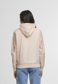 Ženska z dolgimi temnimi lasmi, oblečena v svetlo bež jopico s kapuco in modre cargo kavbojke, obrnjena stran od enostavne podlage.