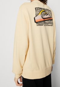 Sudadera de color crema con un corte holgado, que presenta un diseño gráfico en la parte trasera en naranja, negro y gris, con puños acanalados.