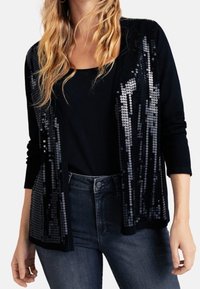 Cardigan nero con dettagli di paillettes lungo il davanti, maniche lunghe, design aperto, indossato sopra una maglietta nera e abbinato a jeans scuri.
