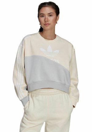 Kort sweatshirt i ljusbeige tyg med grå detaljer, strukturerade ränder och en framträdande vit logotyp på bröstet. Kombinera med matchande byxor.