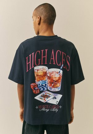 Osoba w czarnej koszulce z napisem "HIGH ACES", na plecach nadrukowane dwa napoje z lodem, żetony do pokera, kości oraz karty do gry.