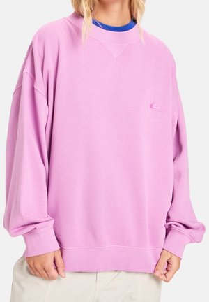 Persoon draagt een oversized lichtpaarse crewneck sweater over een blauw shirt en lichte broek, houdt de zoom van de trui met beide handen vast.