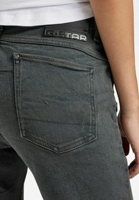Jeans de mezclilla gris oscuro con una textura sutil, logotipo de G-Star en la cinturilla y un bolsillo trasero con detalles de costura.