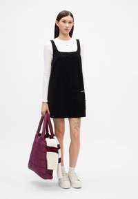 Μαύρο βελούδινο φόρεμα με τετράγωνη λαιμόκοψη, φορεμένο πάνω από ένα λευκό πουκάμισο με μακριά μανίκια. Κουβαλώντας μια μωβ τσάντα με κατασκευή από ύφασμα με καπιτονέ σχέδιο και μια ριγέ μαντίλα κρεμασμένη επάνω.