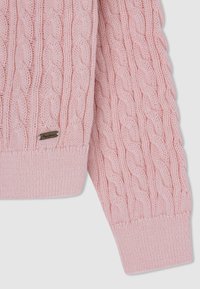 Różowy sweter z dzianiny z wzorem w warkocze, z prążkowanymi mankietami i dołem, z metalową metką na dolnym lewym boku.