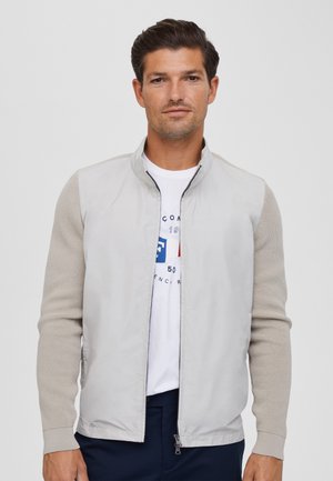 Giacca bomber grigia chiara con maniche a maglia, zip frontale e colletto. La camicia bianca sottostante presenta una grafica blu e rossa.