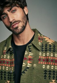 Veste en laine verte à motifs tribaux rouges, noirs et crème, dotée d'un col et d'une fermeture à pression à l'avant, superposée à une chemise noire.