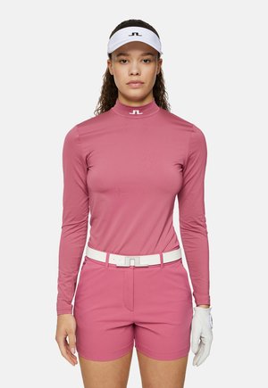 Rosa Langarm-Golfshirt mit hohem Kragen, kombiniert mit passenden rosa Shorts. Wei�ßer Gürtel und Visier mit Logo. Weißer Golfhandschuh an der linken Hand.