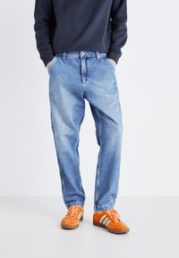 Light blue denim jeans feature a relaxed fit, five-pocket design, and a faded texture. Paired with bright orange athletic shoes.