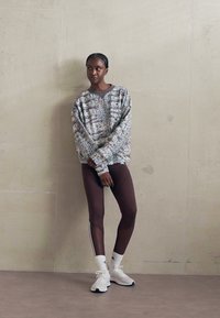 Genser med mønstrete stoff i dempede farger, kombinert med brune leggings med sidestriper og hvite sportssko. Glatte teksturer.