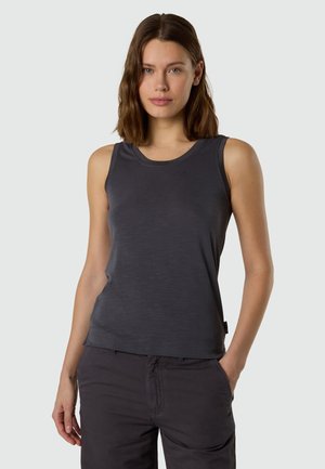 Donna con capelli castani fino alle spalle, indossa una maglia senza maniche grigia scura e pantaloni scuri abbinati, in piedi con una mano in tasca.