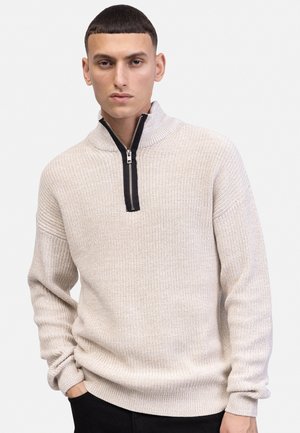 Νεαρός άνδρας που φοράει μπεζ πλεκτό πουλόβερ με μαύρο φερμουάρ στη μέση, στέκεται με το ένα χέρι στην τσέπη μπροστά σε απλό φωτεινό φόντο.