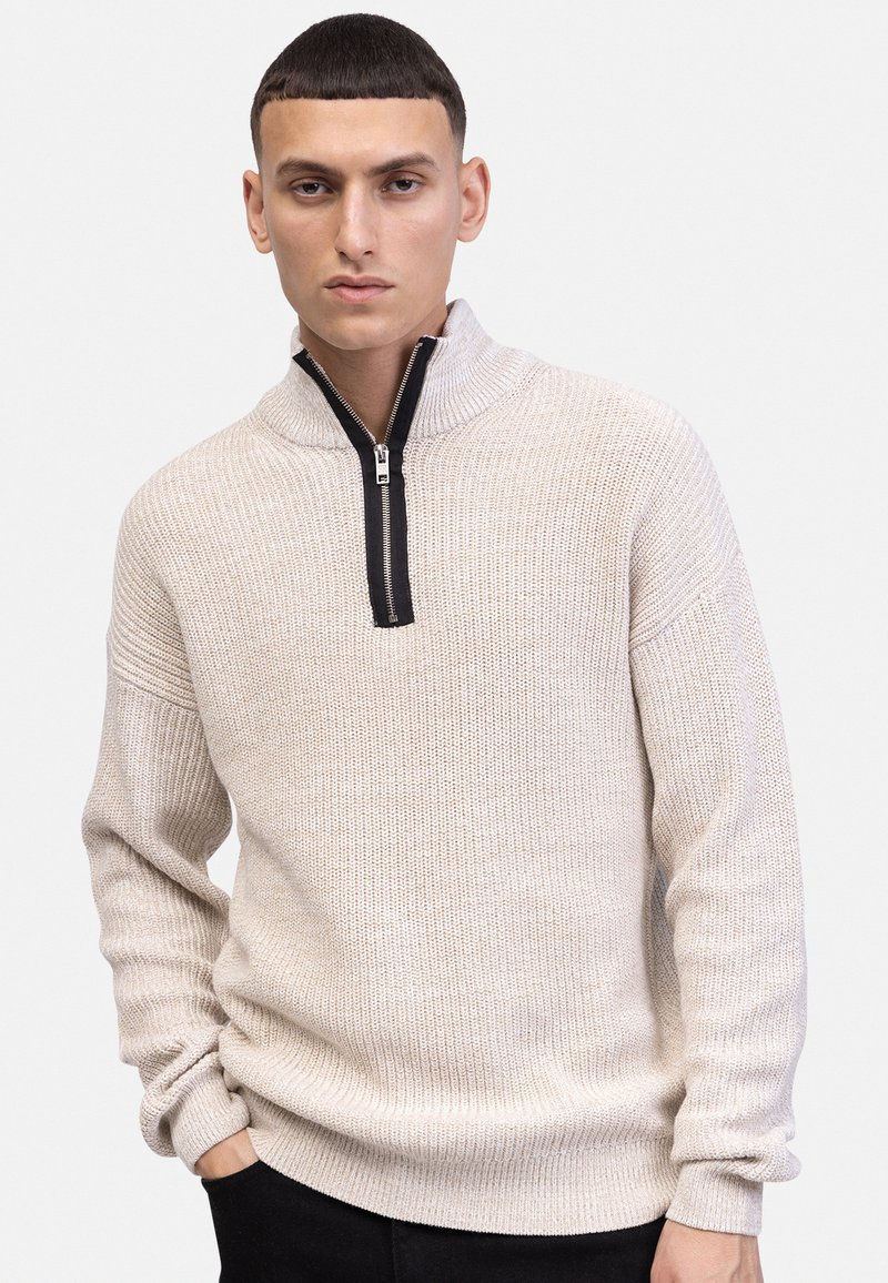 Νεαρός άνδρας που φοράει μπεζ πλεκτό πουλόβερ με μαύρο φερμουάρ στη μέση, στέκεται με το ένα χέρι στην τσέπη μπροστά σε απλό φωτεινό φόντο.