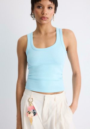 Donna con top azzurro senza maniche e pantaloni color crema con portachiavi a forma di cono gelato, in piedi davanti a uno sfondo chiaro uniforme.