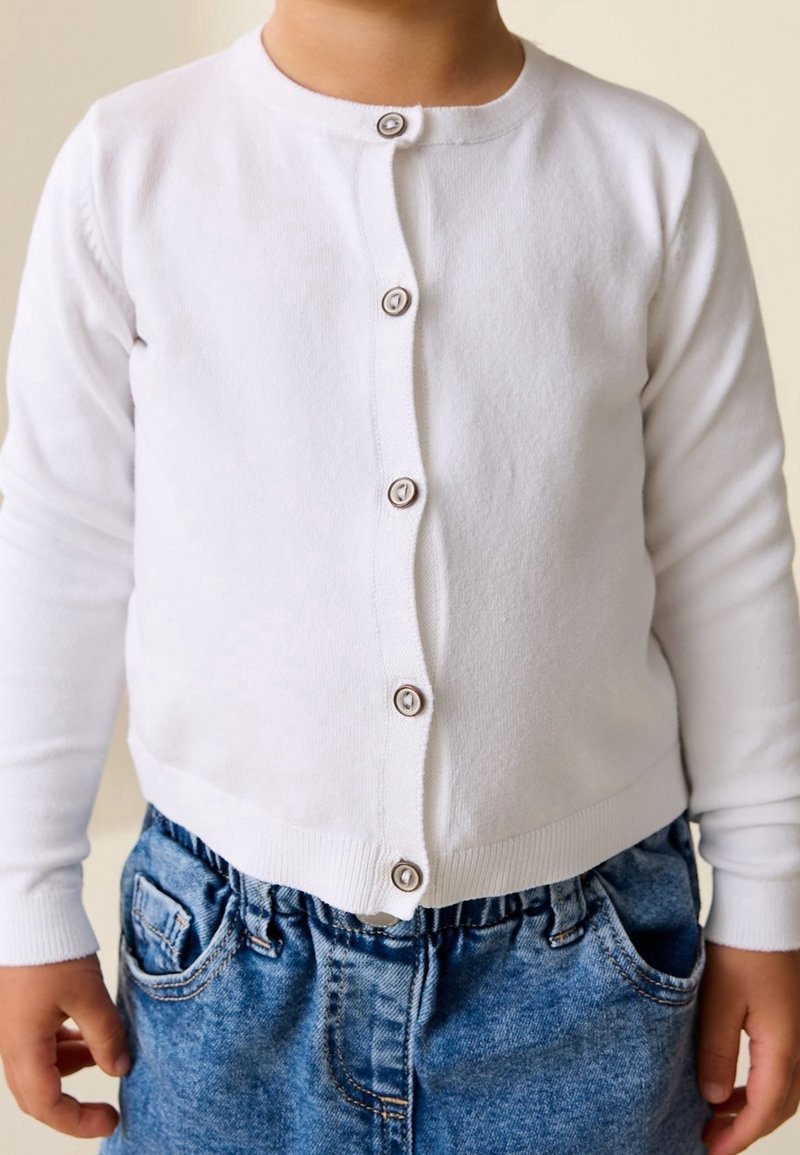 Enfant portant un cardigan blanc boutonné et un jean bleu en denim, mains détendues le long du corps, fond neutre.