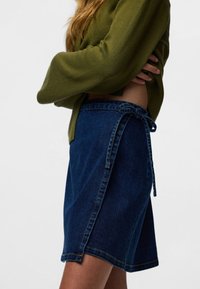 Krilo iz jeansa v temno modri barvi z zavezovanjem ob strani. Tkanina ima gladko teksturo z nežnimi šivi ob robu.