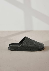 Mustavi mustasta kudottua nahkaa, suljettukärkinen ja litteapohjainen, beige tekstuuri pinnalla pehmeillä varjoilla.