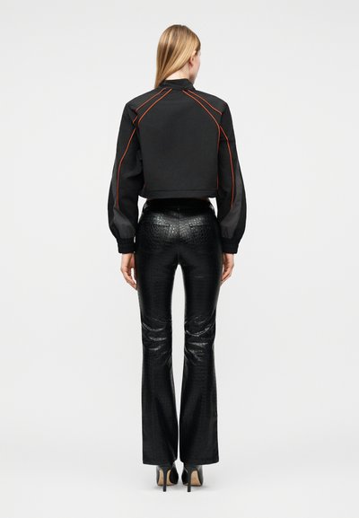 Γυναίκα που φορά μαύρο κοντό μπουφάν με πορτοκαλί διακοσμητικές ραφές και μαύρο υφασμάτινο ψηλόμεσο παντελόνι καμπάνα, στέκεται πλάτη σε μονόχρωμο φόντο.