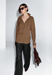 Brown oversized shirt featuring a collar, paired with dark wide-leg jeans and a brown handbag with studded accents. Glossy dark sunglasses worn.
