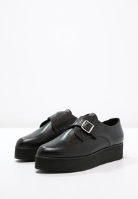 Chaussures à plateforme en cuir noire avec un bout rond, présentant un accent en boucle latérale et une semelle épaisse et texturée. Fini lisse partout.