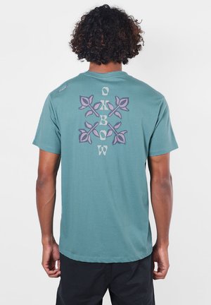 Homme aux cheveux bouclés portant une chemise turquoise à manches courtes avec un motif floral violet et un texte au dos, debout devant un fond uni.