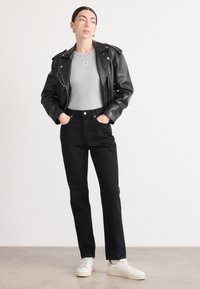 Donna che indossa una giacca di pelle nera, una maglietta grigia chiara, jeans neri a vita alta e scarpe da ginnastica bianche, in piedi con le mani nelle tasche su uno sfondo bianco.